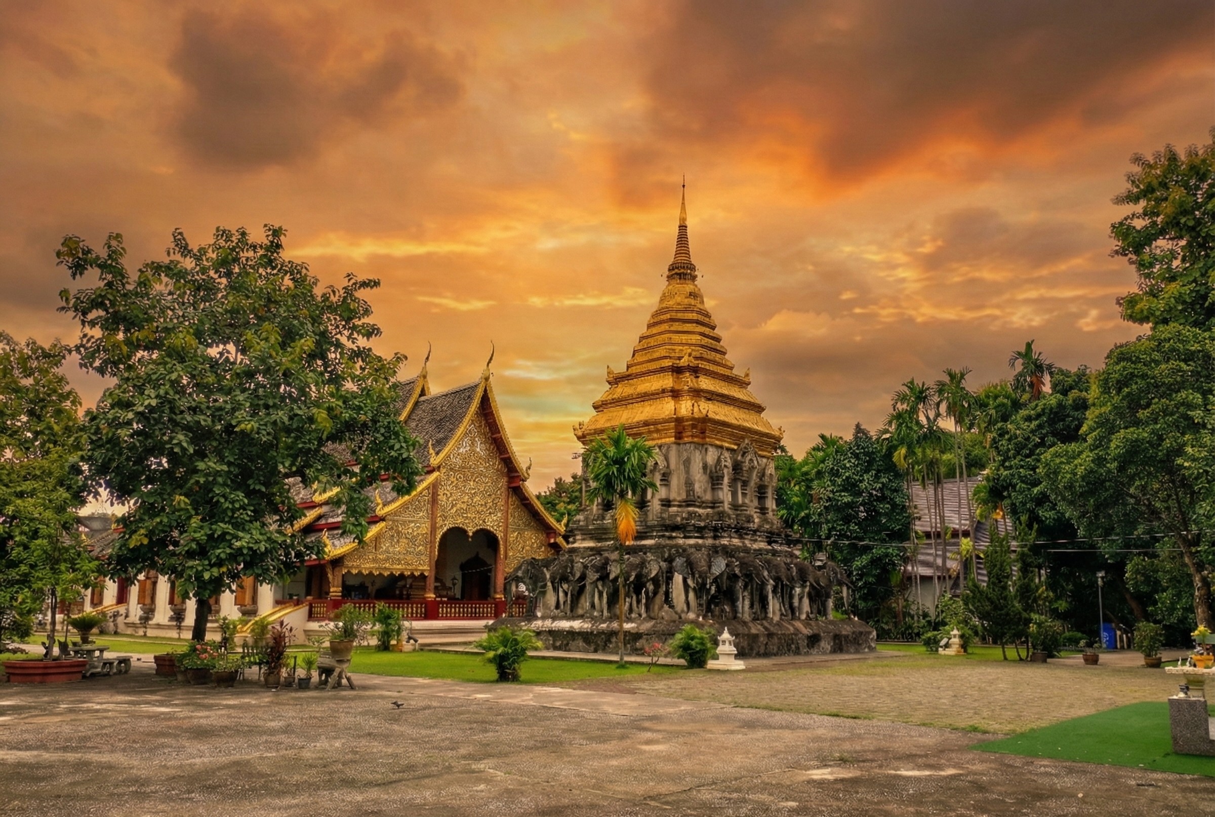 Golden sunset over Wat Chiang Man, Chiang Mai's oldest temple, featuring the prominent Chedi Chang Kham, or Elephant Chedi, adorned with elephant statues, and the Lanna-style Viharn in the foreground under a dramatic sky.