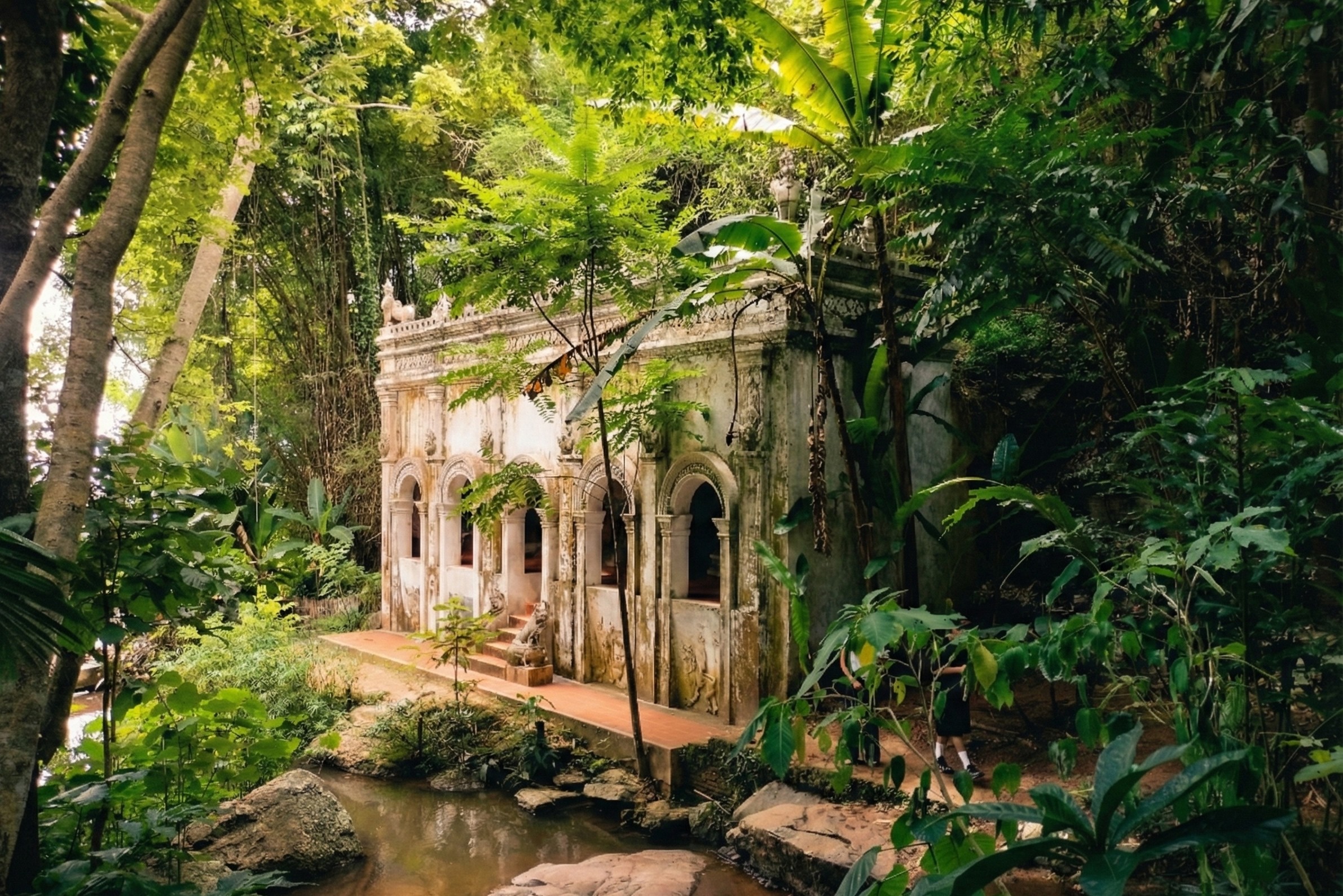 Ancient white masonry pavilion featuring Burmese-style arches and weathered stucco, nestled within the lush jungle greenery at Wat Pha Lat in Chiang Mai, with a natural stream flowing over rocks in the foreground.